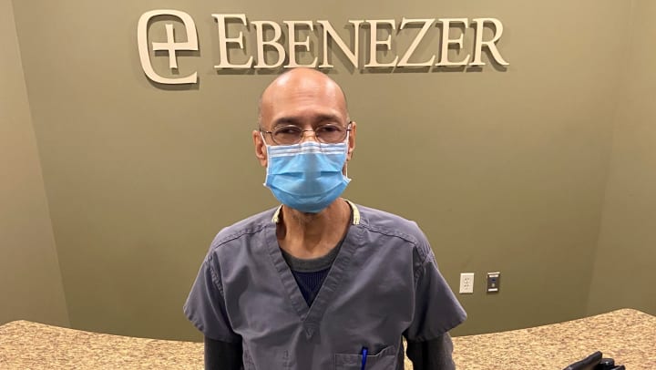 Ebenezer Nursing Assistant Zabir “Zee” Ali wearing scrubs and a face mask and standing in front of an Ebenezer sign and welcome desk.