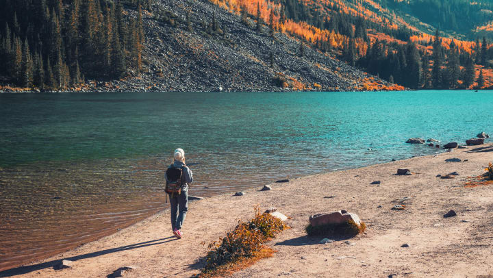 A person stands in front a body of water with mountains and fall foliage in the distance