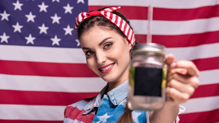  Woman with a red and white striped headband standing in front of American flag holding a Mason jar beverage holder with straw toward the camera. 