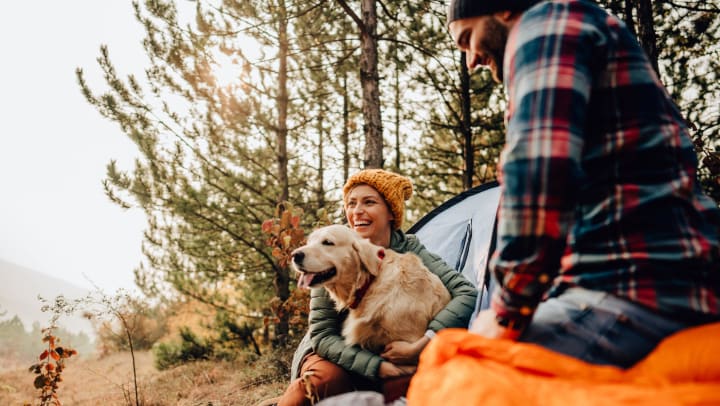 Man and woman outdoors surrounded by camping equipment, the woman is hugging a golden retriever.