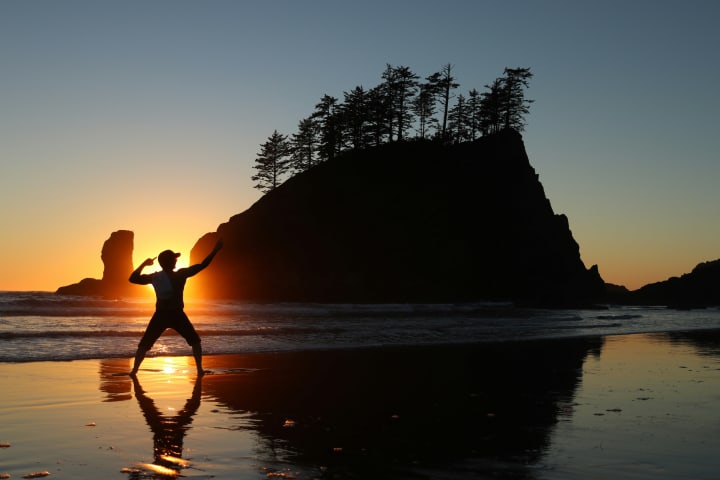 beach scene at sunset with someone standing in front of a large land form in the ocean at second beach in la push Washington