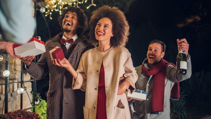 Two warmly dressed men and a woman smiling and holding gift boxes and a bottle of champagne, are on the front porch of a home decorated with holiday lights. 