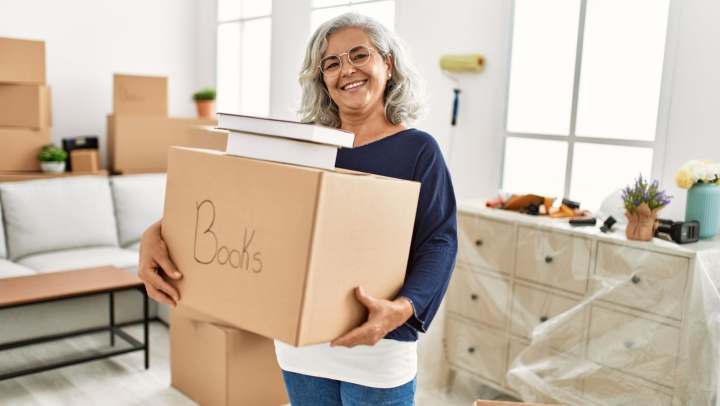 A smiling woman holds a box in a room filled with moving supplies 