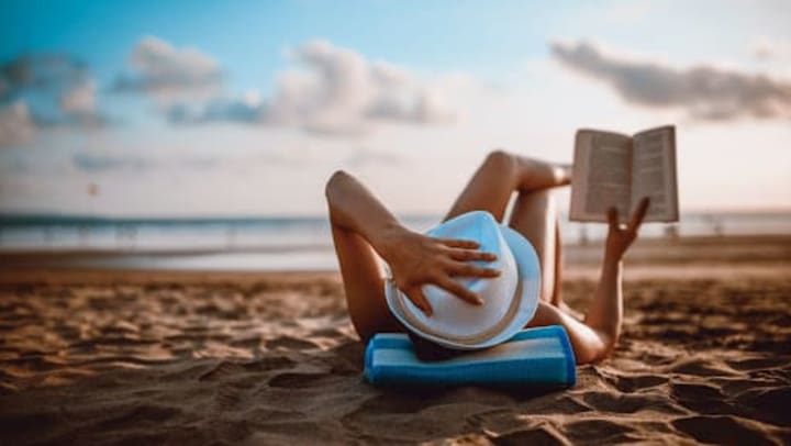 A woman lounging on the beach with a hat on holding a book up as she reads.