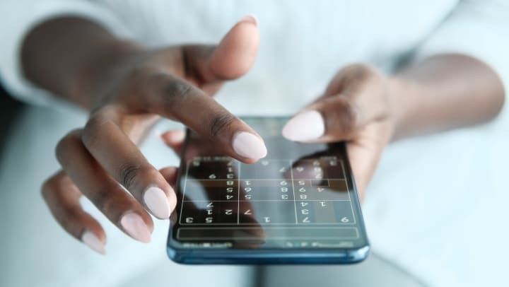 A person is wearing a white outfit, using both of their hands, painted with light pink nail polish is holding an iPhone, playing a game of Soduku 