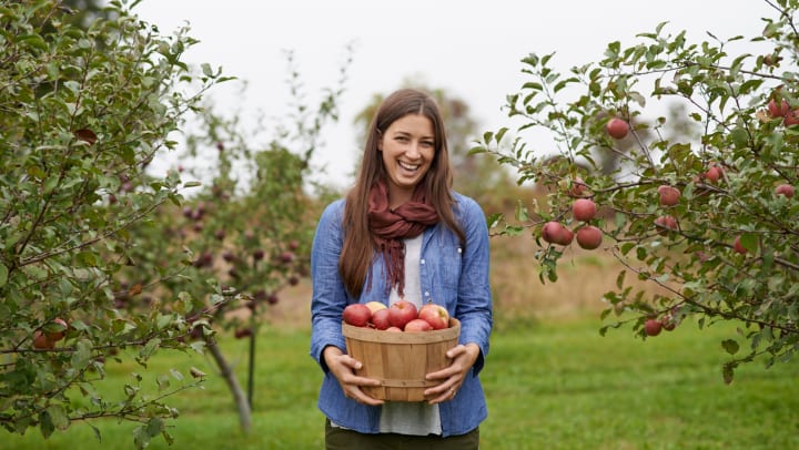Woman standing between apple trees, smiling while holding a wooden bucket full of red apples.