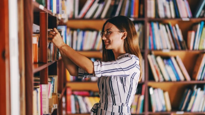 Young woman in glasses standing next to bookshelves and searching for a book | bookstores in Pompano Beach
