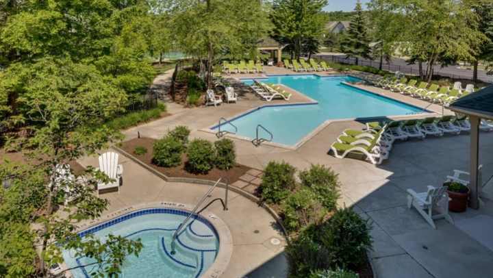 Green lounge chairs resting near a circular hot tub and a large blue swimming pool reflecting the sky near tall trees.