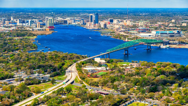 Aerial view of the beautiful St. Johns River cutting through Jacksonville with green trees in the foreground and the Hart Bridge crossing the river