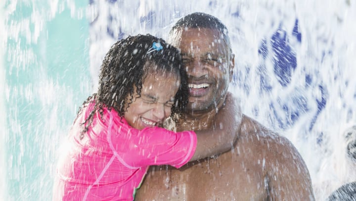 Father and daughter smiling and holding each other as water falls on them at a water park