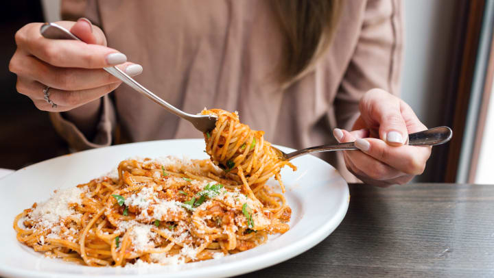 Woman using a fork and spoon to eat plate of spaghetti.