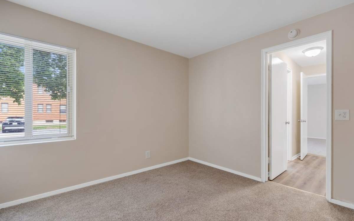 Bright carpeted apartment bedroom with window and hallway access at Highland Manor Apartments in Rochester, New York