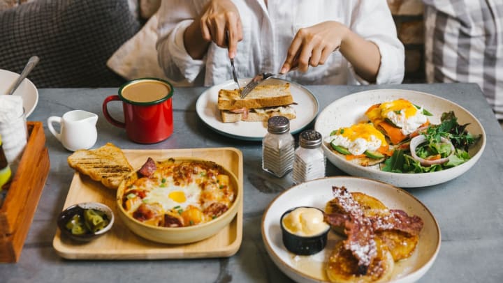 A person uses a knife and fork to cut into a breakfast sandwich at a table also showing three other plates of breakfast foods | breakfast spots in Charlotte