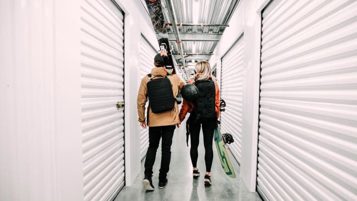 A man and woman walking through a self storage corridor, each holding assorted outdoor gear