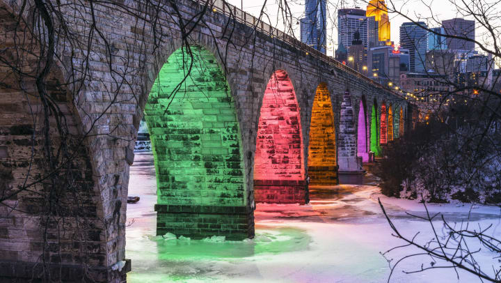 Stone arch bridge over iced water with holiday lights with the Minneapolis skyline behind it.