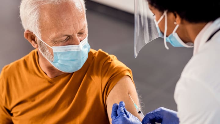 An older man wearing a face mask receiving a vaccine injection in his upper arm. 
