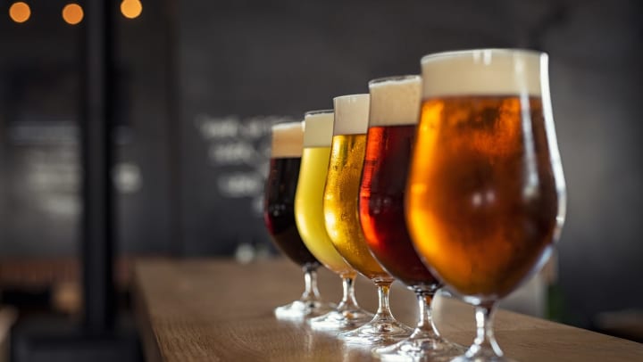 Different colored beers in glasses set on top of a bar with an out of focus industrial background.