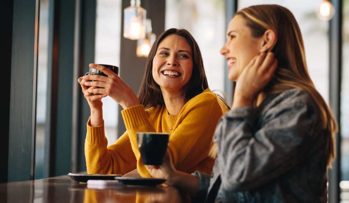 Two women enjoying coffee and laughing at a modern café with warm lighting at Valley Brook Townhomes in Rochester, New York