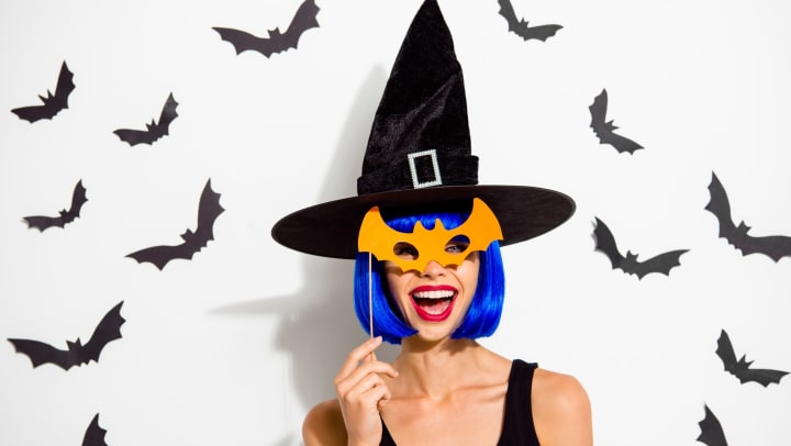 Woman in a blue wig wearing a black pointed hat and holding an orange bat mask up to her face. 