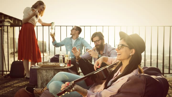 Young woman sitting on a balcony and playing the guitar, while three other young adults chat and hang out in the background.