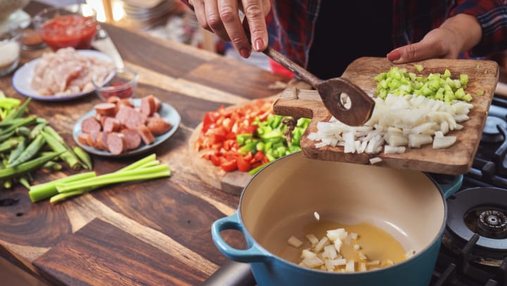 Person holding a cutting board and pushing chopped onions and celery into a Dutch oven on a stovetop. Sliced sausage, chicken, okra, and red bell peppers can be seen in the background.