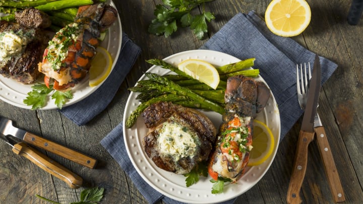 Lobster, steak, and asparagus on a plate with utensils and lemon wedge set on a wood table.