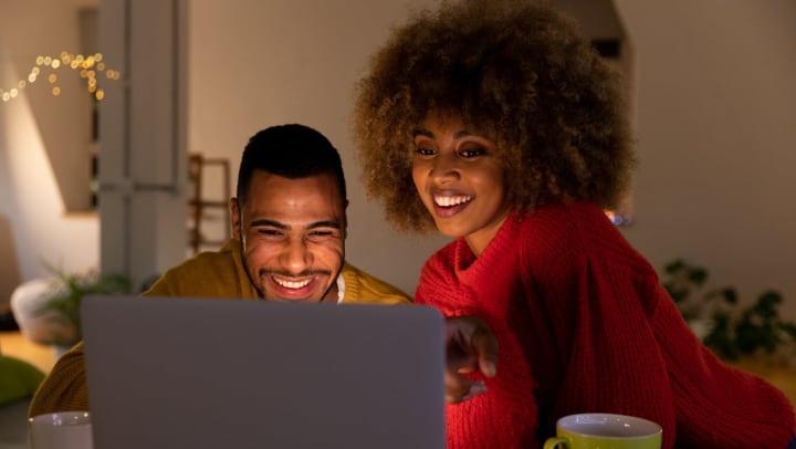A smiling man and woman sitting at a table with coffee mugs nearby, looking at a laptop computer together.