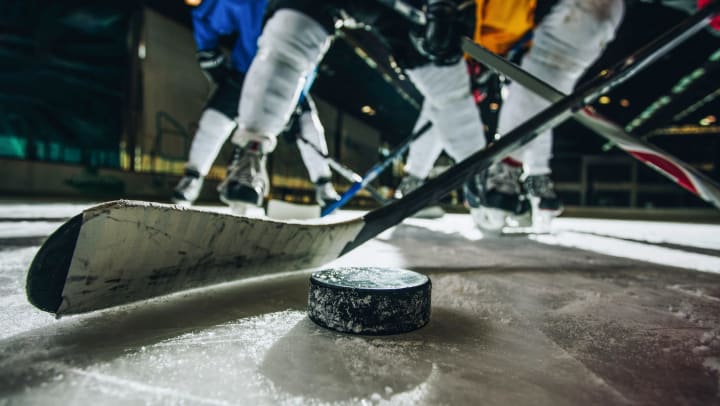Close-up of a hockey stick about to strike a puck. Hockey players are seen in the background.