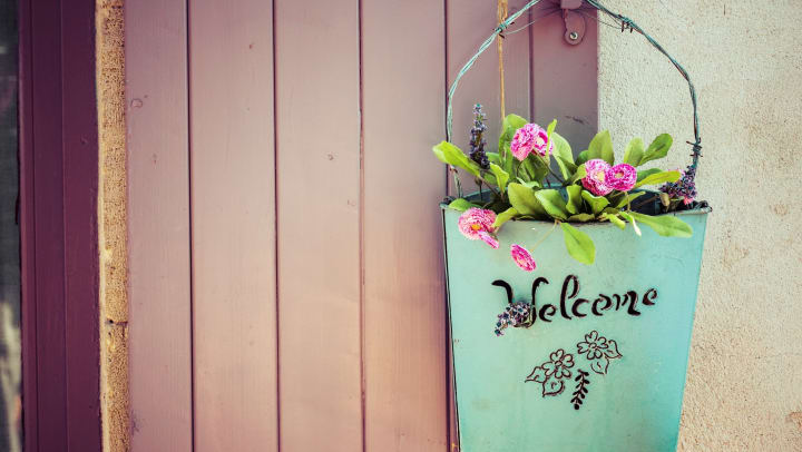 A basket hung on a purple door that reads “Welcome” and holds pink flowers.
