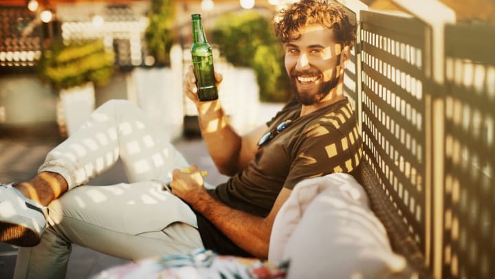 Man sitting outdoors, leaning against lattice, with a beer in his hand, smiling at the camera.