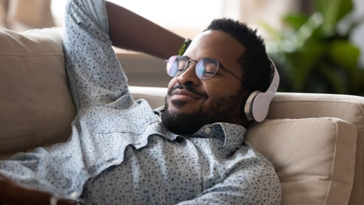 Man wearing glasses with headphones on while lying on the couch with a smile on his face.