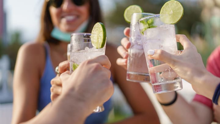 People toasting four glasses containing a clear liquid and ice, garnished with a lime wheel.