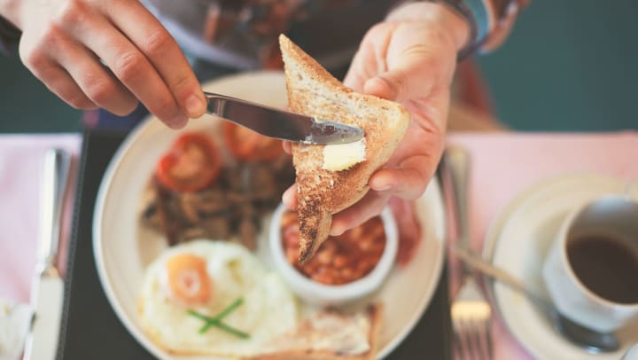 Closeup of a person’s hands buttering toast with a plate of breakfast food on the table in the background.