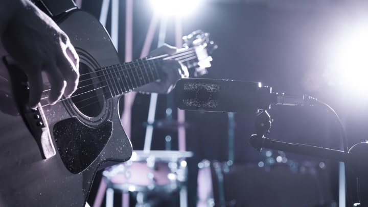 Closeup of microphone recording an acoustic guitar in a recording studio or concert hall, with a drum set in the background out of focus.