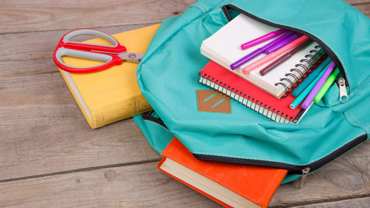 A light blue backpack on its side with a bunch of supplies falling out including notebooks, scissors, books, and colored markers.