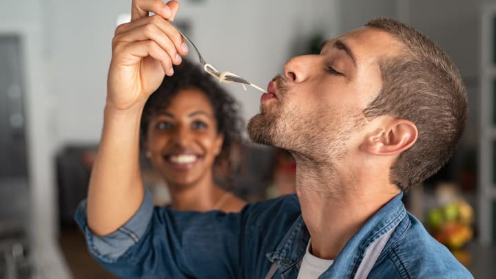 A man putting noodles in his mouth with his fork while a slightly out of focus smiling woman looks at him.