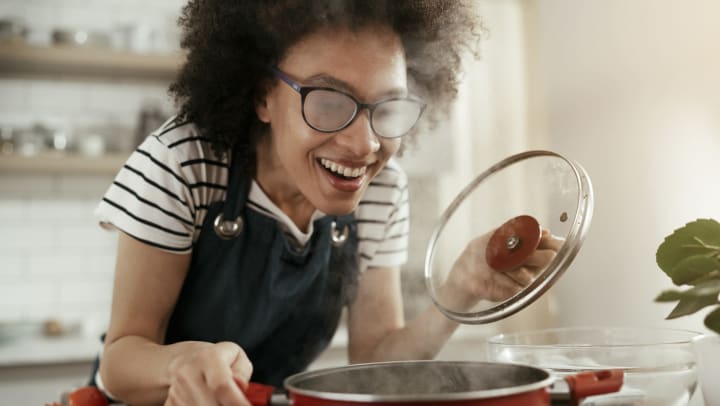 A smiling woman leaning over a red pot with steam billowing up from it. 