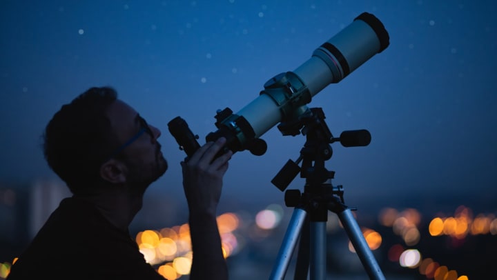 Man with his hand on a telescope pointed at the night’s sky, with city lights behind him. 