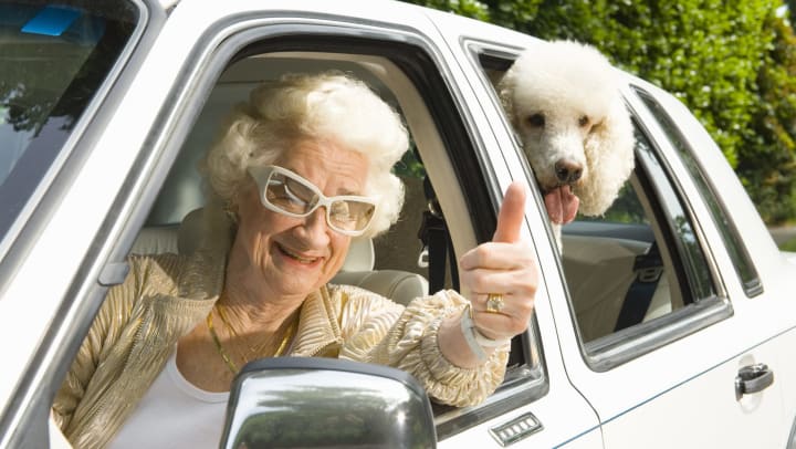 A smiling senior gives the thumbs up from behind the wheel of a car, while a dog looks out the rear window.