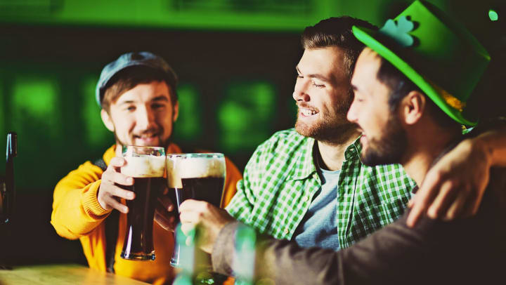 Three friends enjoying dark beer, clinking tall glasses at counter of Irish pub on St, Patrick’s Day