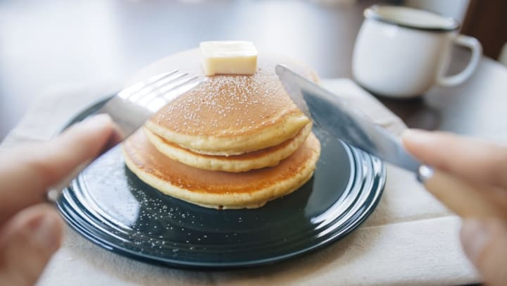 A person holding a knife and fork that is about to dig into a stack of pancakes with butter on top | pancakes near Keller