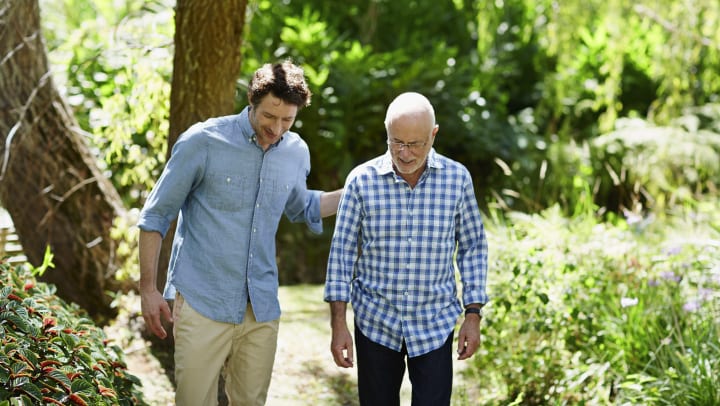 A younger man with his hand on the back of an older man who both have a smile on their face and are walking outside.