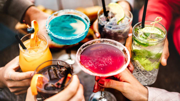 Six people toast a variety of glasses containing a mix of colorful cocktails, including margaritas, a gin and tonic, and more