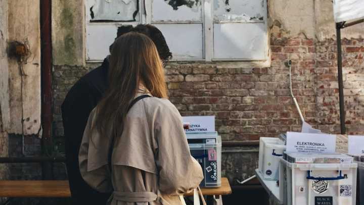 A man and a woman stand with their backs to the camera, looking down at items on a table at an outdoor market or fair. The woman wears a tan trench coat. They are standing against a background of an old brick wall with a dilapidated, white-framed window above. The table in the foreground holds several white plastic bins.