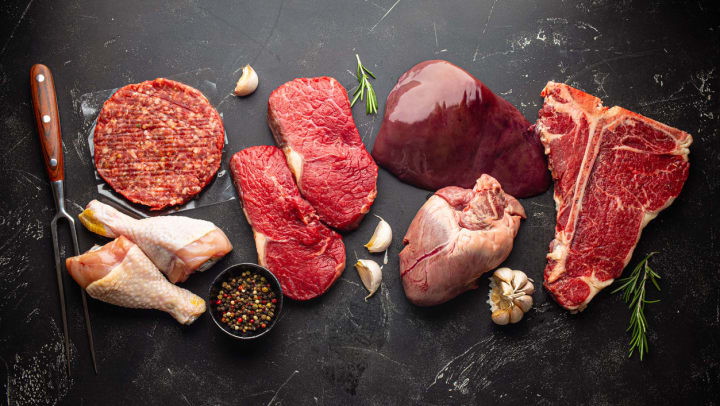 A selection of uncooked beef steak, ground meat patty, heart, liver and chicken legs on black stone background from above.