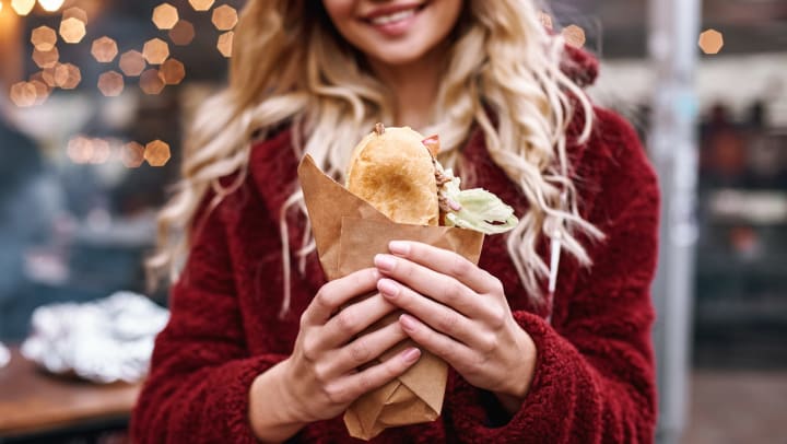 Woman in a red sweater holding a sandwich wrapped in parchment paper in front of her. 