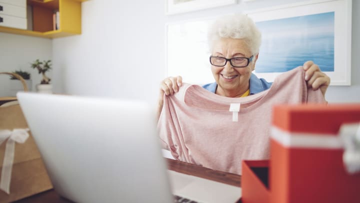 Senior woman holding up a shirt and smiling with an open box and open laptop computer in the foreground.