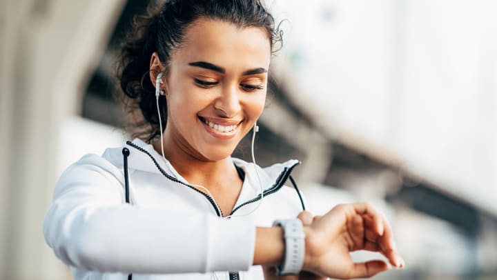 Smiling young woman wearing headphones and a light jacket looking at the face of her wristwatch