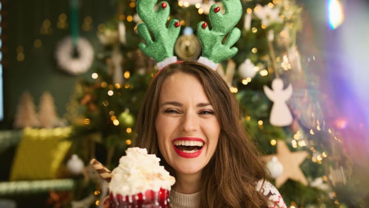 Woman wearing Christmas tree ears, standing in front of a Christmas tree and holding a drink with whipped cream, smiling at the camera.