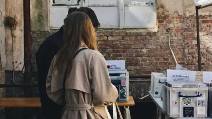 A man and a woman stand with their backs to the camera, looking down at items on a table at an outdoor market or fair. The woman wears a tan trench coat. They are standing against a background of an old brick wall with a dilapidated, white-framed window above. The table in the foreground holds several white plastic bins.